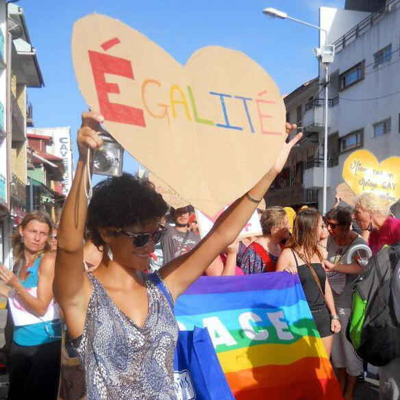 Katherine standing, waist up - short dark curly hair, with sunglasses, blue tank top, and holding heart sign with egalite written out in rainbow letters